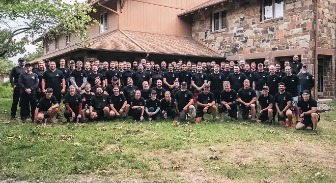 Group photo of about 60 smiling men in black shirts, posing outside a large stone and wood house. They stand on green grass, exuding camaraderie and joy.