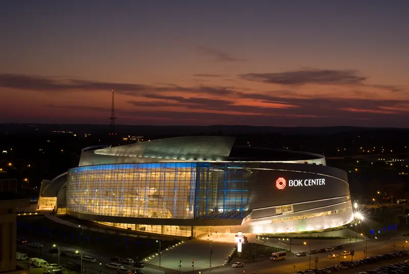 A modern arena, the BOK Center, is illuminated in blue at dusk, set against a vibrant sunset sky. The scene is calm and architecturally striking.