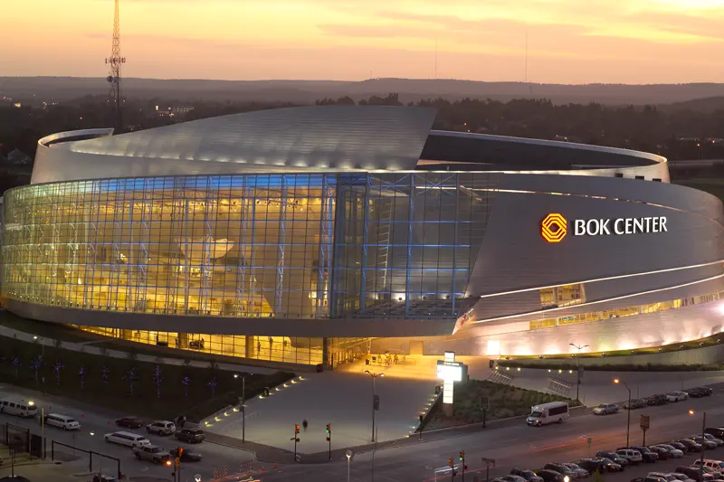 Modern arena, BOK Center, glows warmly against a sunset sky. Its sleek, curved glass facade is illuminated, conveying a vibrant, bustling atmosphere.