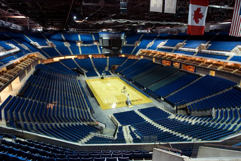 Spacious indoor basketball arena with empty blue seats surrounding the central wooden court. Canadian flag hangs prominently, evoking a calm, expectant atmosphere.