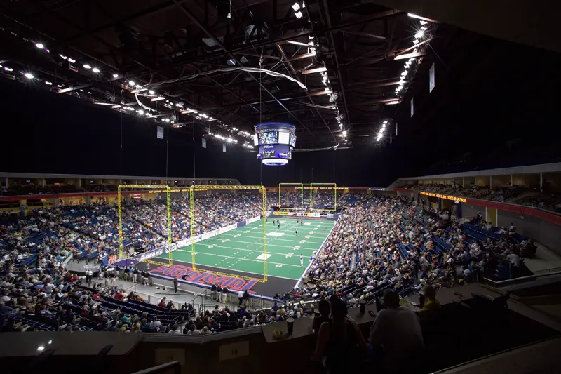 A packed indoor football arena with a green field, yellow uprights, and a large crowd. The atmosphere is lively and bright, highlighting the sports event.