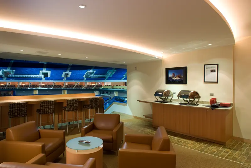 Luxurious stadium suite with tan leather chairs, a glass coffee table, and a view of the brightly lit blue-seated arena. Buffet with chafing dishes on the side.