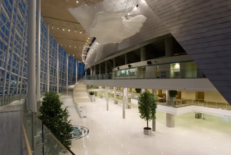 Spacious modern atrium with high ceilings, large windows, and sleek design. White floors, suspended art piece, and potted trees add elegance.