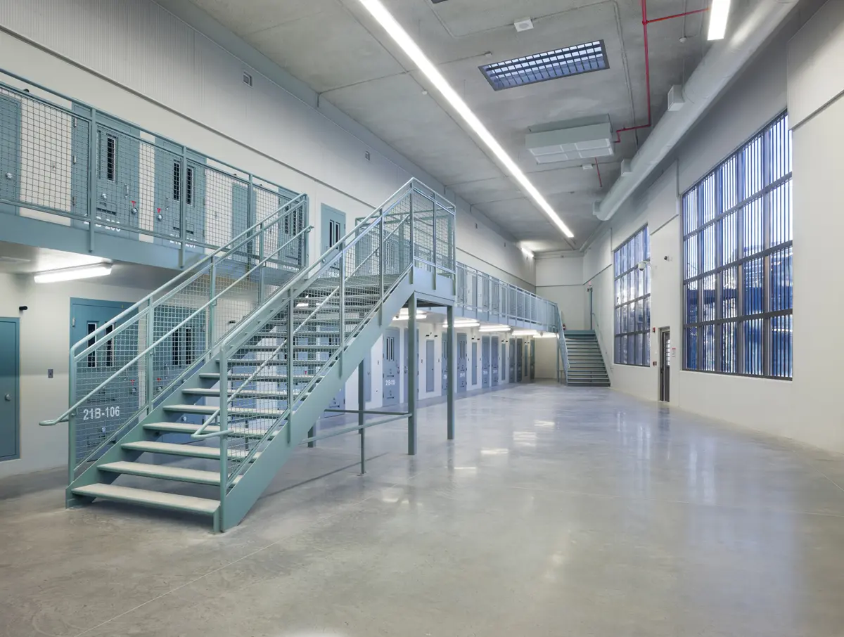 Industrial-style interior of a large, empty hall with metal stairs and upper walkway, flanked by cell doors. Harsh lighting creates a cold, sterile tone.