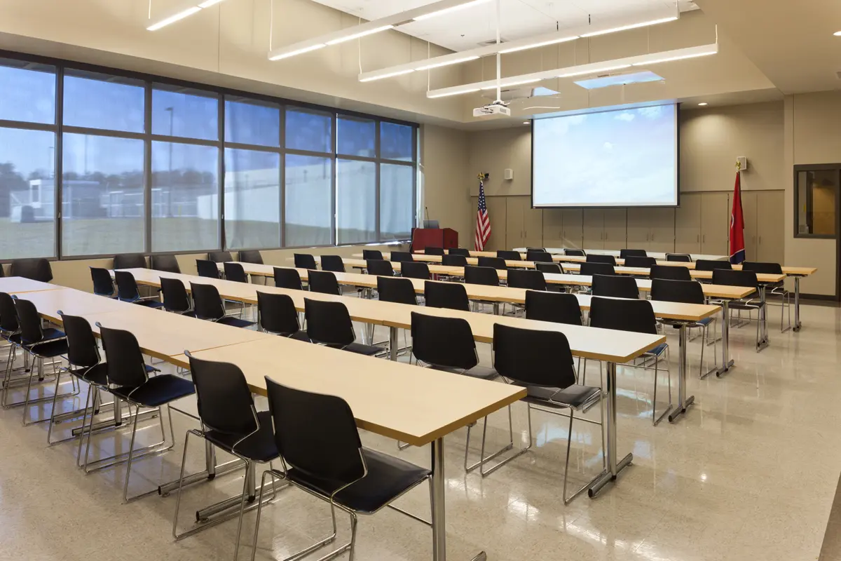 A spacious, modern conference room with rows of long tables and black chairs, a large screen, American flags, and bright windows along one wall.