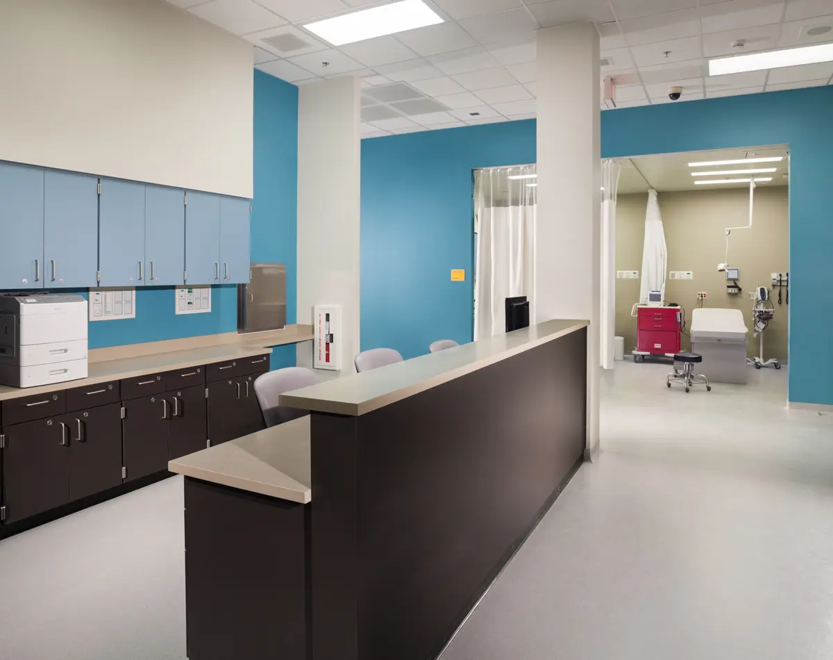 A clean, modern hospital nurse station with a dark counter and blue walls. Cabinets with medical documents are above. A patient room with equipment is visible beyond.