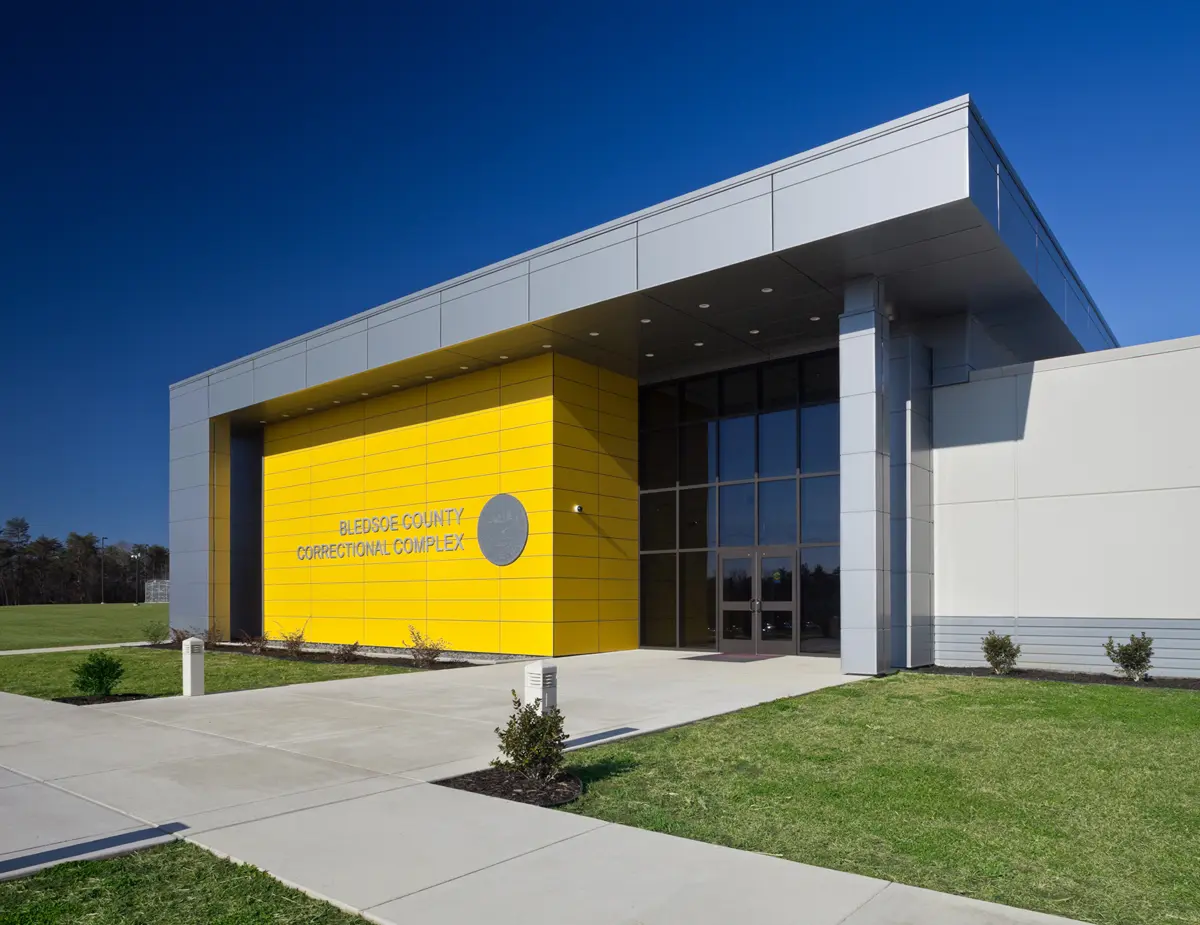 Modern correctional complex entrance with a bright yellow accent wall, grey facade, large glass doors, and green lawn under a clear blue sky.