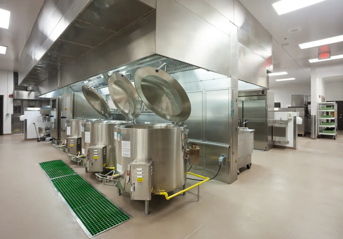 A sterile, industrial kitchen features large stainless steel kettles with open lids under a metal vent hood. The space is clean and well-lit.