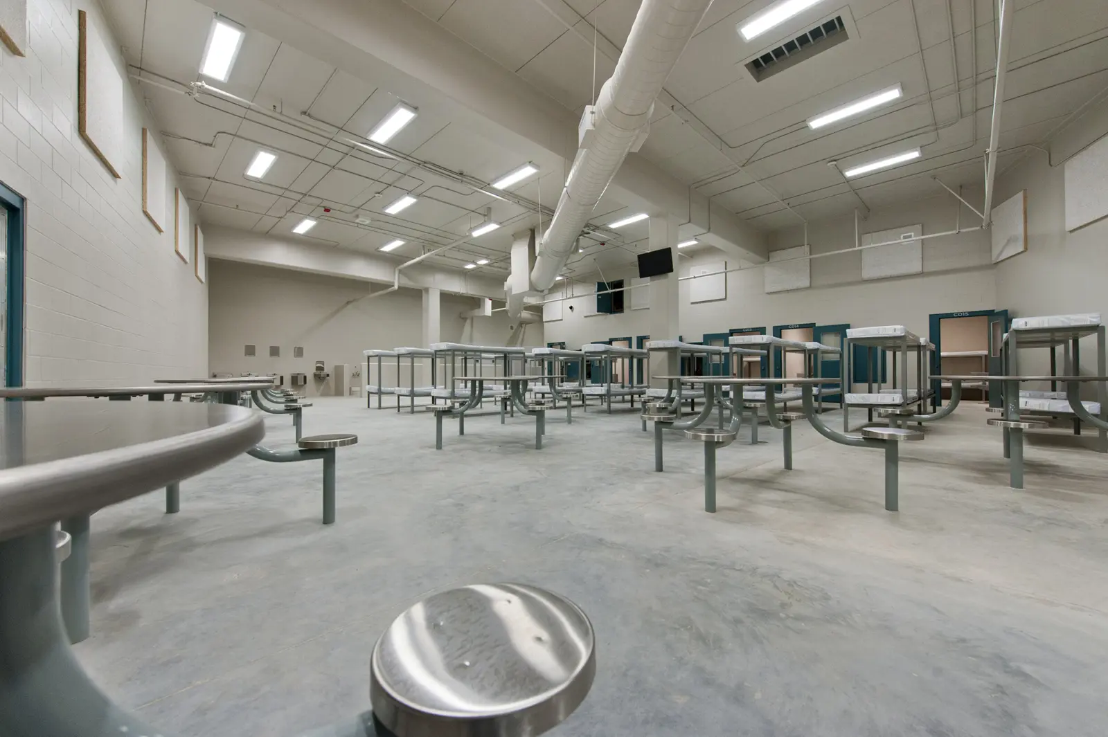 Spacious, sterile room with metal tables and benches under bright fluorescent lights. Industrial ceiling ducts enhance the cold, institutional atmosphere.