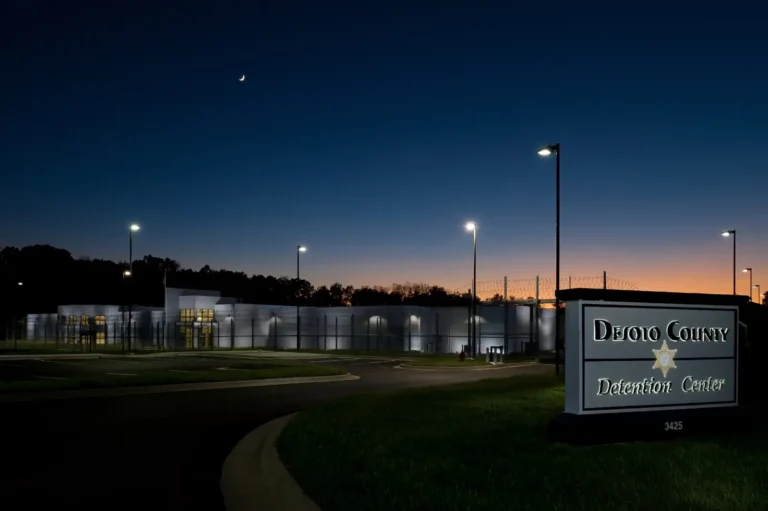 Night view of a detention center with bright street lamps, a crescent moon in the sky, and an illuminated sign reading "DeSoto County Detention Center."