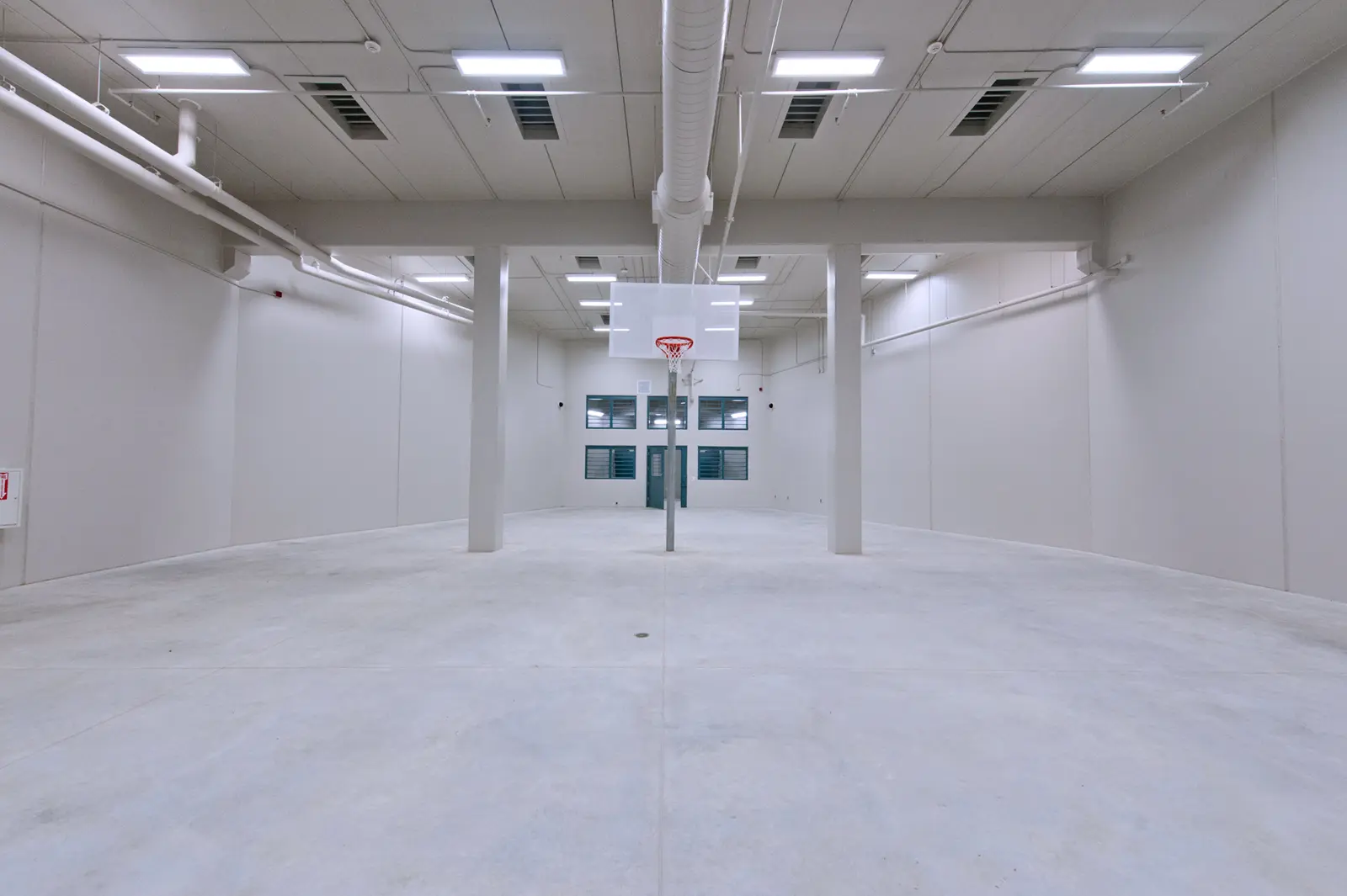 Spacious, empty indoor room with high ceilings, minimalistic white walls, and a central basketball hoop. Bright lighting adds a stark, sterile feel.