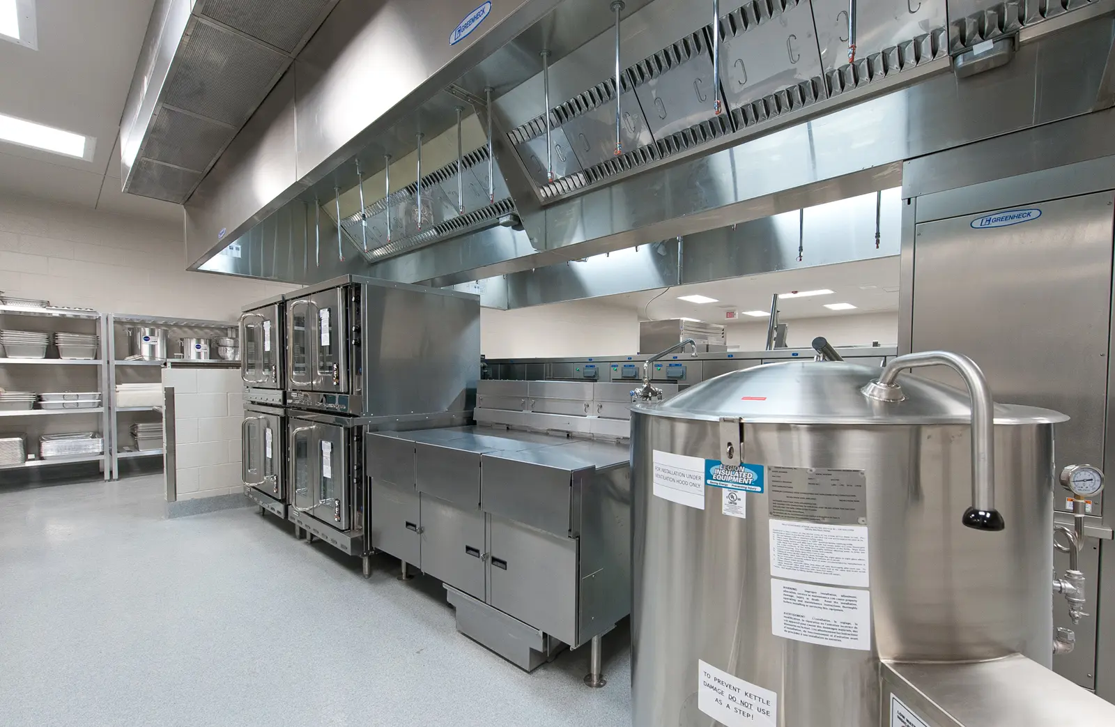 Commercial kitchen with shiny stainless-steel appliances, large ovens, and storage racks. The space is modern, clean, and brightly lit, conveying efficiency.