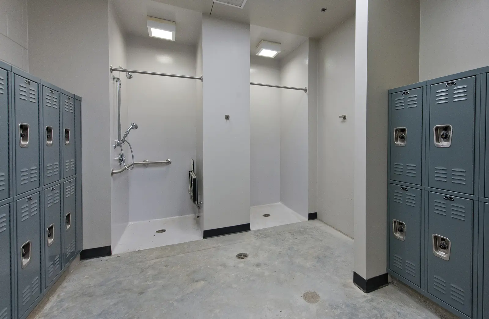 Locker room with two shower stalls and gray lockers. The showers have grab bars and a minimalist design. The space feels clean and utilitarian.