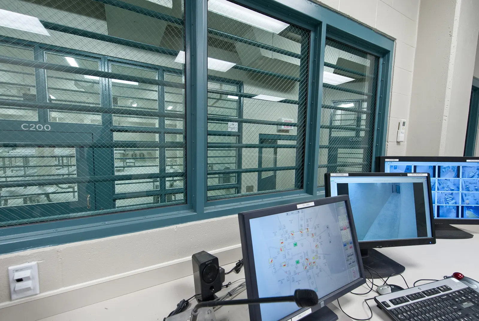 Control room with monitors and a view into a facility through reinforced glass windows. Two screens show surveillance footage, creating a secure atmosphere.