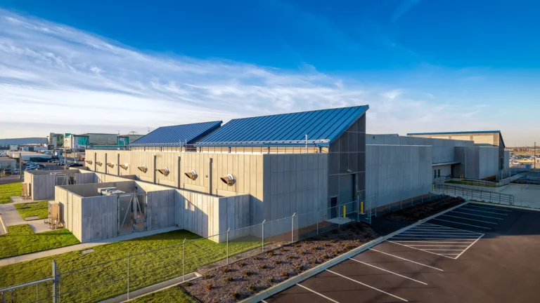 Modern industrial building with a blue roof and gray walls, surrounded by a fence. Empty parking lot and clear sky, creating a calm, orderly atmosphere.