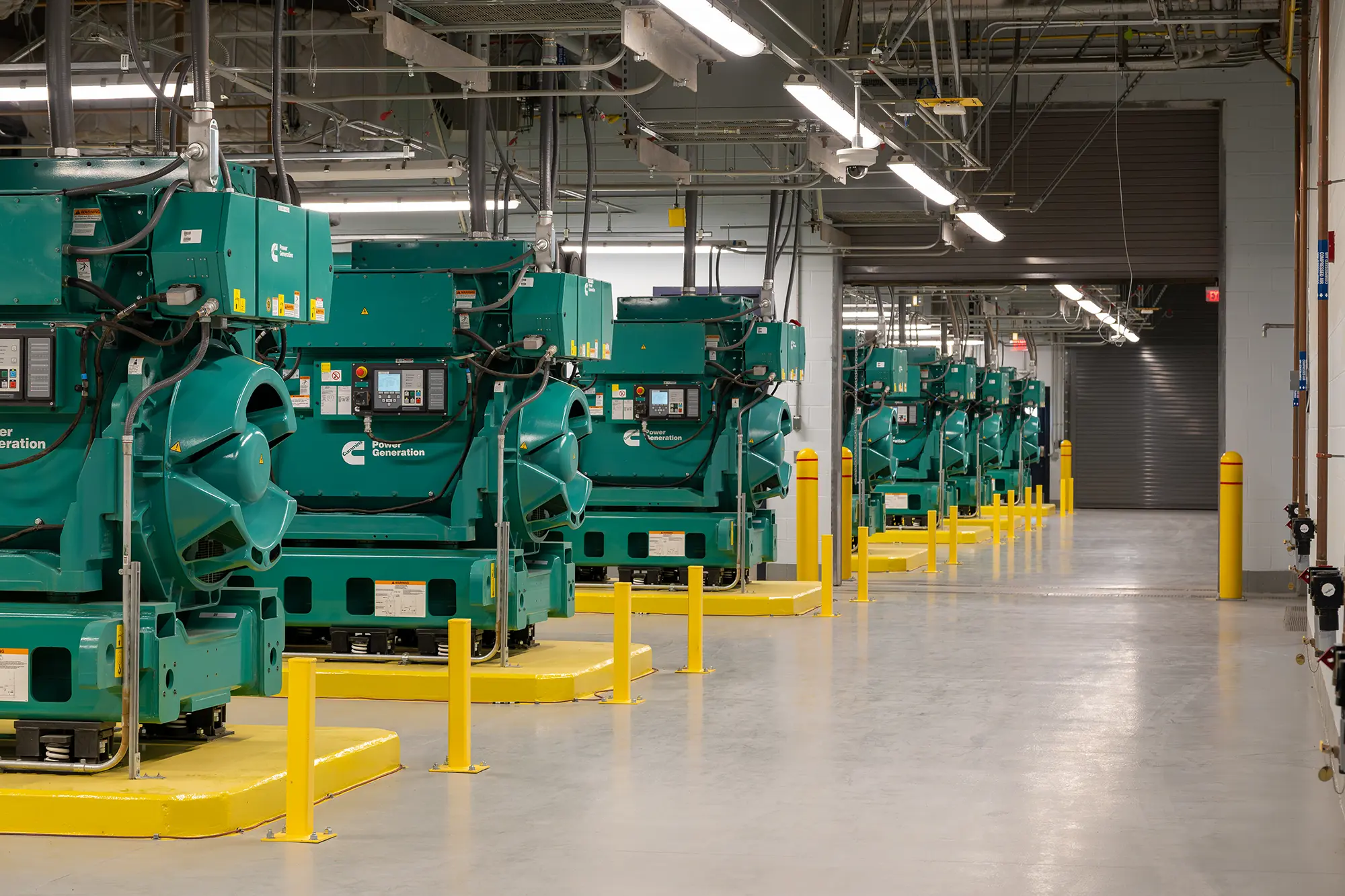 A row of large, green industrial generators lined up in a spacious, well-lit facility. Yellow safety barriers surround each unit. Modern, clean atmosphere.