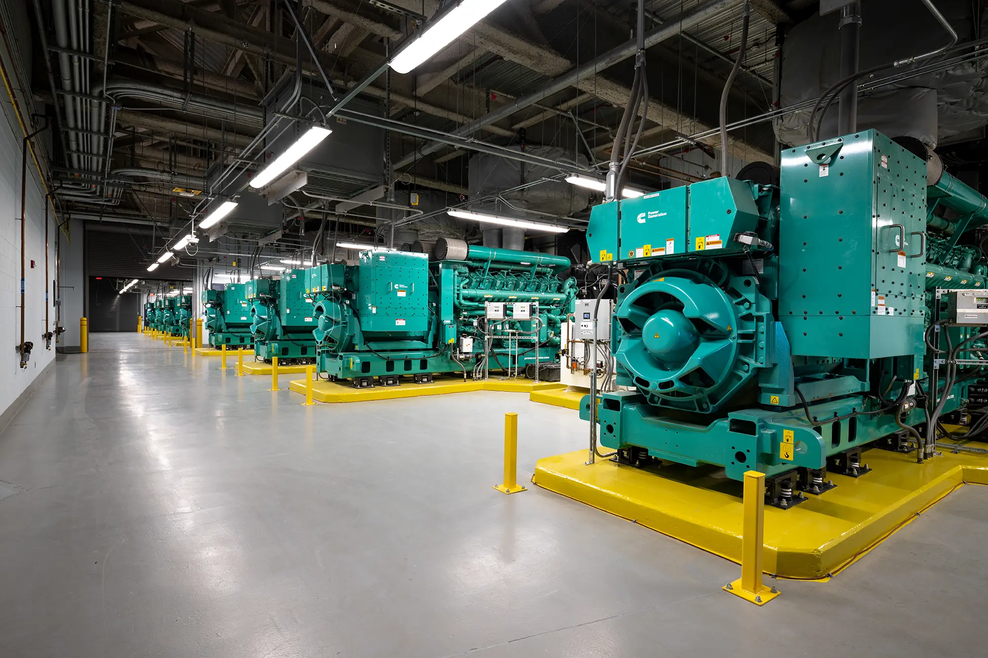 A row of large, teal industrial generators in a well-lit facility with a concrete floor. The generators are on yellow platforms and surrounded by pipes.