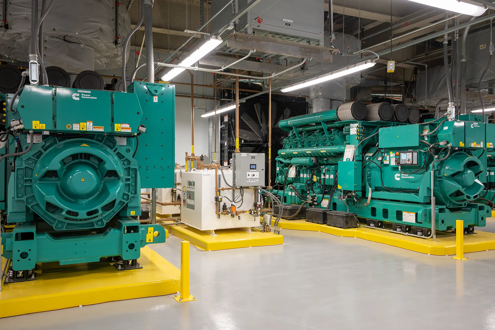 Large green industrial generators in a clean, well-lit room with beige flooring and overhead fluorescent lights. The scene conveys a sense of efficiency.