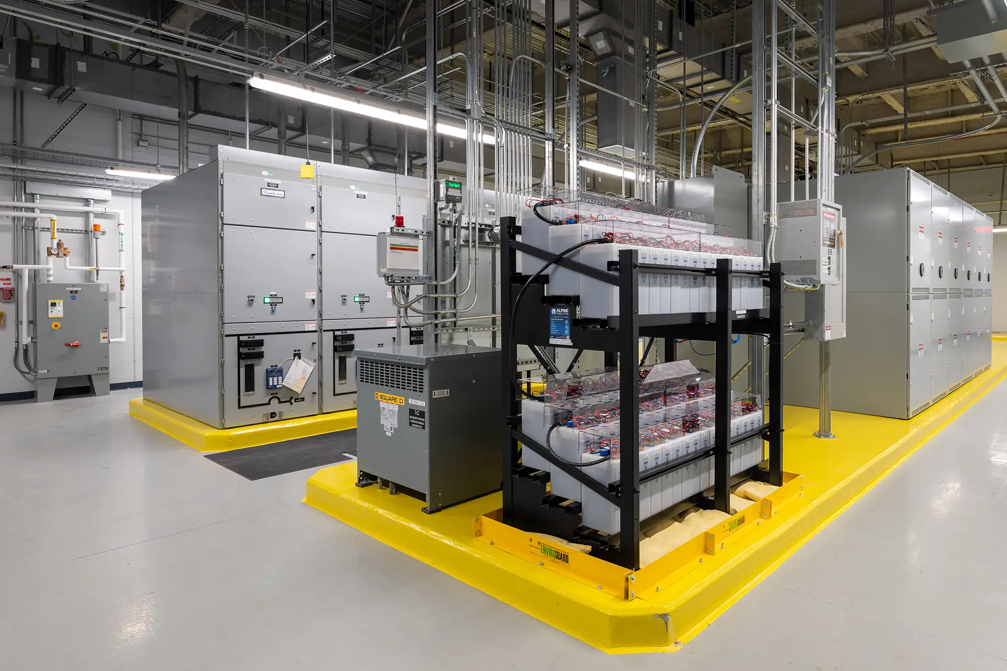 A modern server room with grey server racks, cables, and a backup power system. The clean, industrial setting is highlighted by bright overhead lighting.