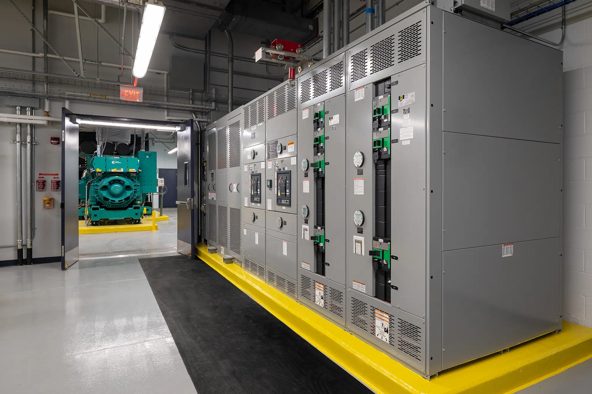 Industrial room with large grey electrical panels and meters on a bright yellow platform. A doorway reveals green machinery in the adjacent room.