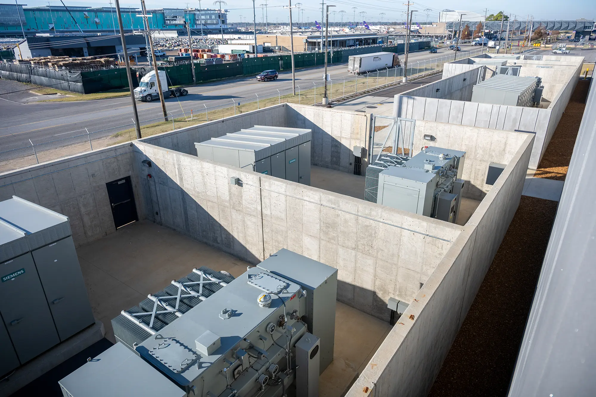 Aerial view of an industrial facility with large transformers enclosed by concrete walls. Roads and trucks are visible in the background under a clear sky.