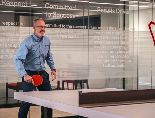 A man in a blue shirt plays ping pong in an office with motivational text on glass walls, creating an atmosphere of focus and enthusiasm.