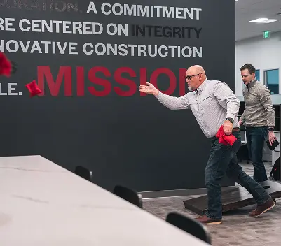 Two men play cornhole indoors, aiming red bags at a target near a wall with motivational text. The atmosphere is focused and playful.