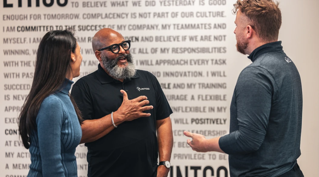 Three people engage in a lively discussion in front of a motivational wall. The atmosphere is positive and collaborative, with two men and one woman smiling.