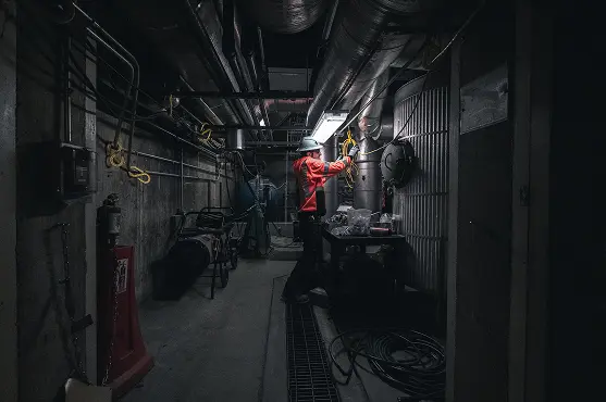 A worker in a red jacket and hard hat, illuminated by a single light, repairs electrical wiring in a dark industrial basement, creating a focused, determined tone.