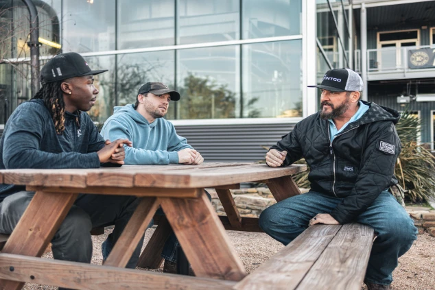 Three men sitting at a wooden picnic table, engaged in conversation. They wear casual outdoor clothing and hats. The setting is relaxed by a modern building.