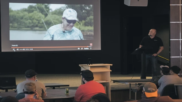 A man presents in a dimly lit room with an audience, showing a projected image of a smiling person fishing. The atmosphere is relaxed and informal.