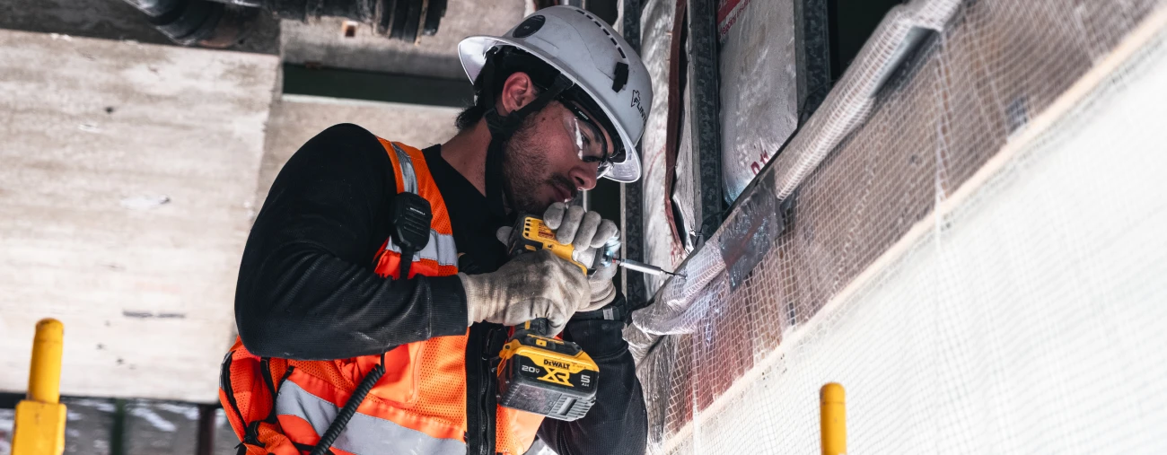 A construction worker in a white hard hat and orange vest uses a power drill on a building site. The scene conveys focus and industry.