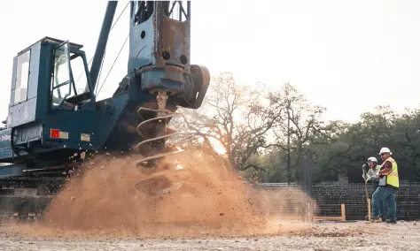 A large drill churns up dirt at a construction site, with dust swirling around. Two construction workers in safety gear observe. Trees in background.
