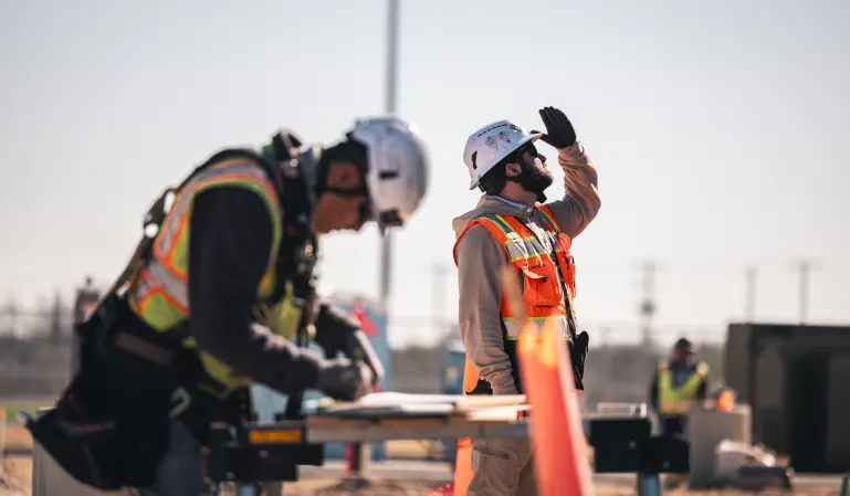 Two construction workers in safety gear are on a sunlit site. One writes on a clipboard; the other shades his eyes, looking upward. A focused, industrious atmosphere prevails.