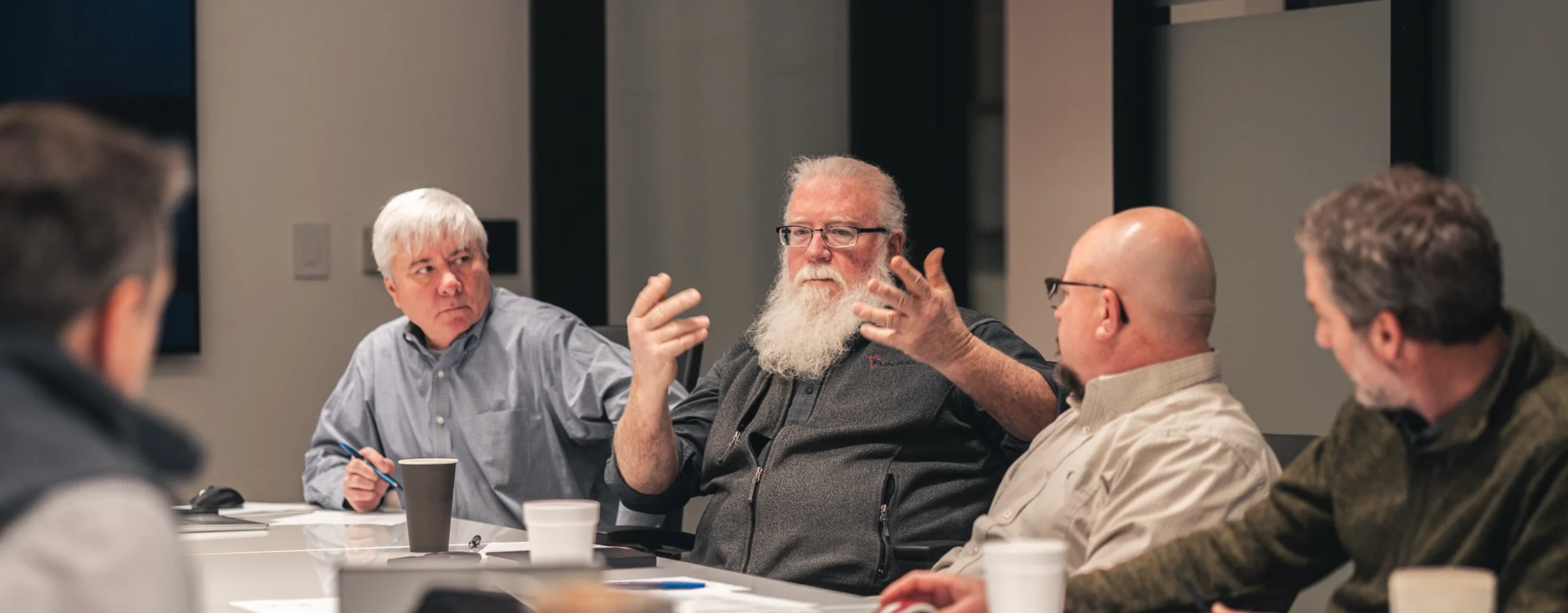 A group of five men having a discussion around a conference table. The central figure gestures passionately, creating an engaged and focused atmosphere.