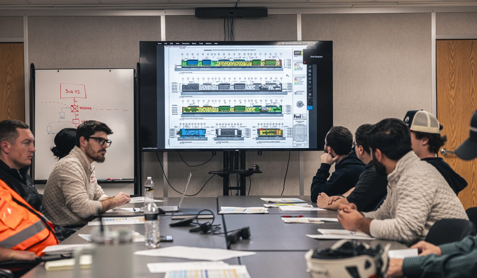 A group of people in a conference room attentively view detailed diagrams on a large screen. A whiteboard with notes and door are in the background.