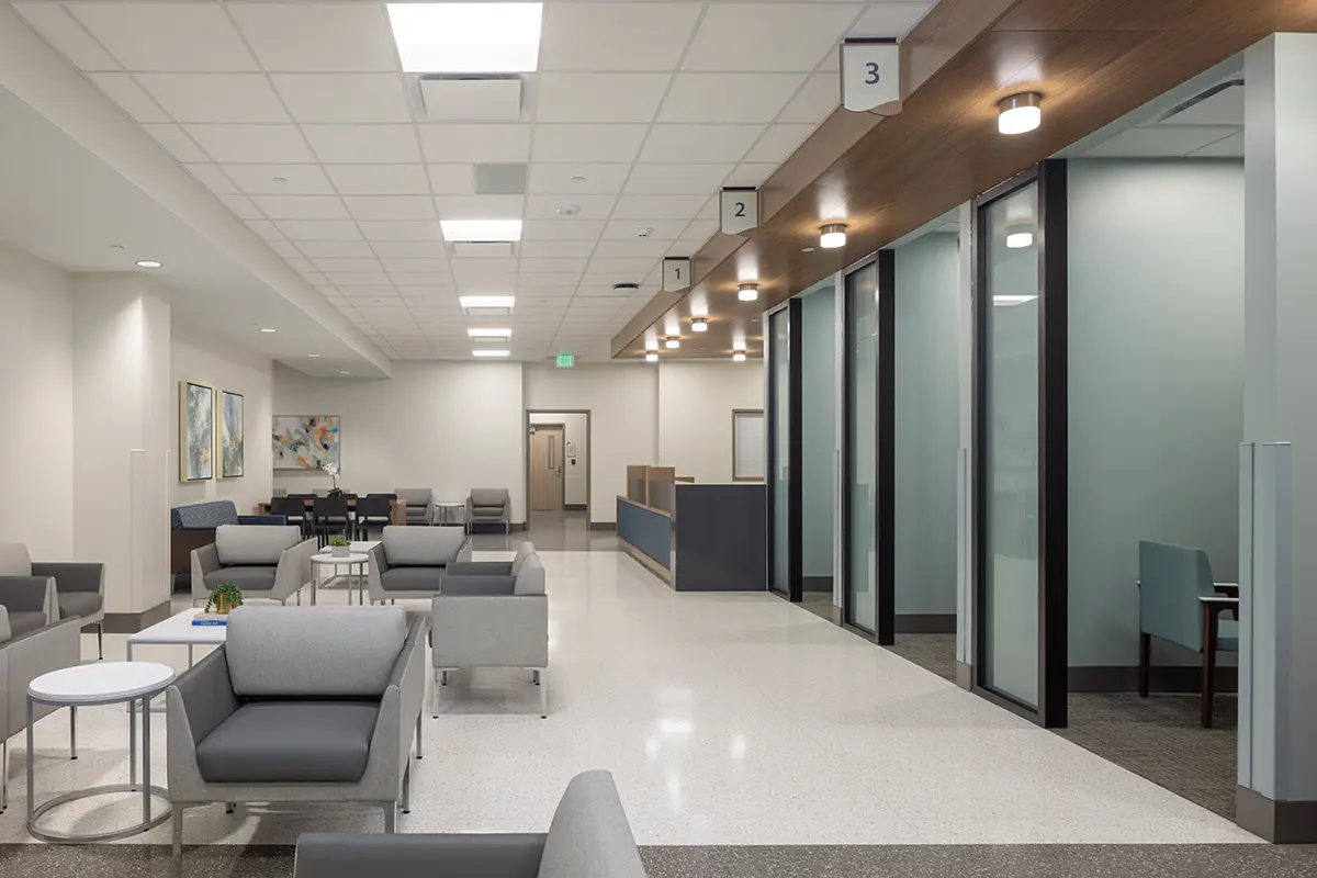 Modern waiting area with gray sofas and round tables in a spacious room. Glass-walled offices on the right, neutral tones, bright lighting. Calm ambiance.
