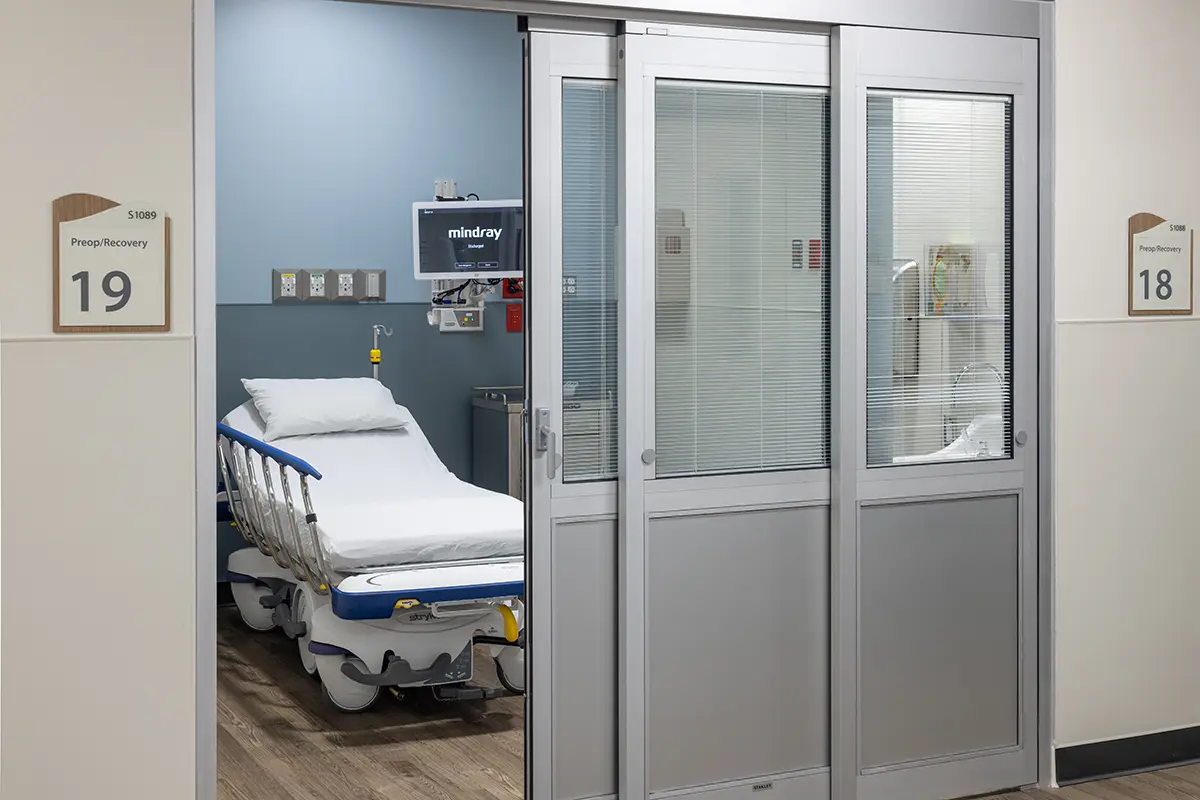 Hospital room with sliding glass doors open to a bed equipped with medical devices on a neat wood floor. Room numbers 18 and 19 are visible.