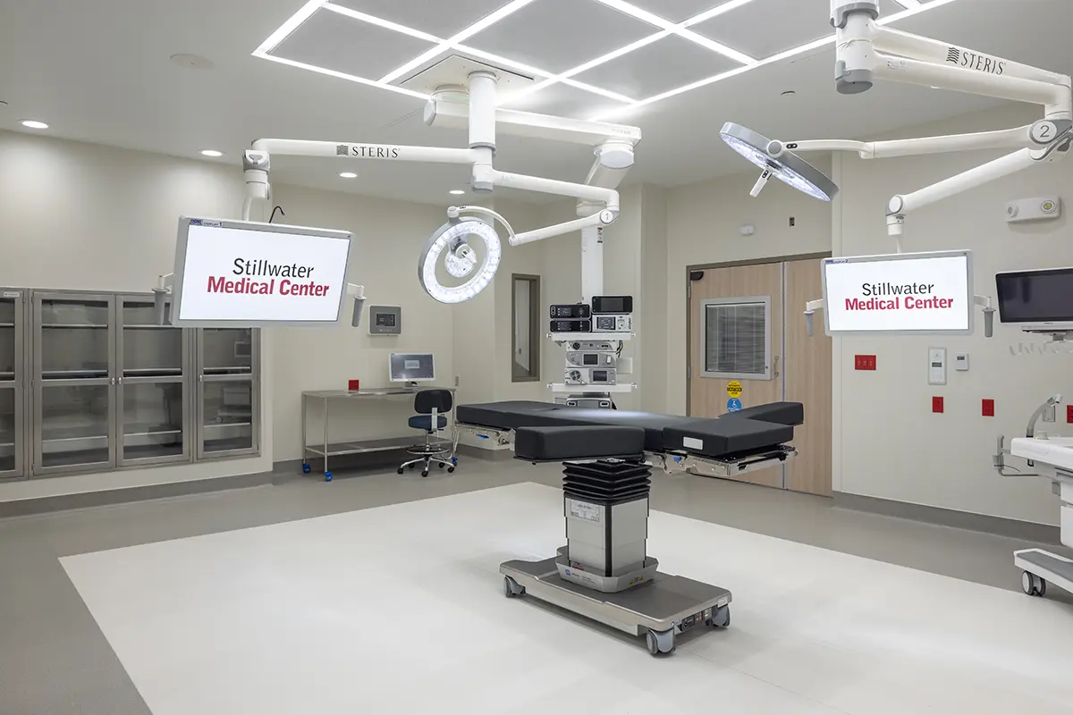 Modern hospital operating room with an adjustable surgical table, overhead lights, and monitors displaying "Stillwater Medical Center." Clean and sterile.