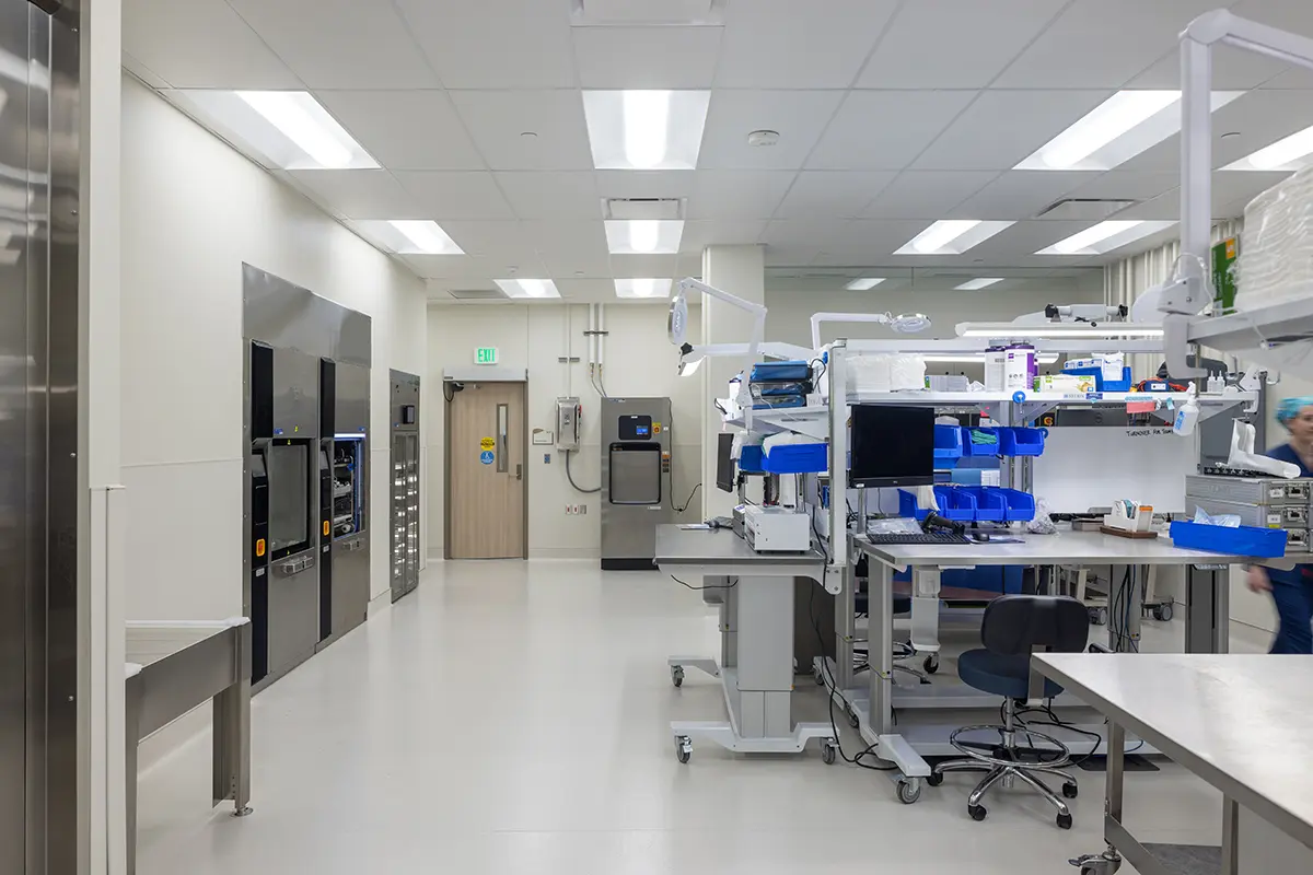 A modern, sterile laboratory with bright lighting, containing workstations with computers and equipment. A technician in scrubs moves at the back.