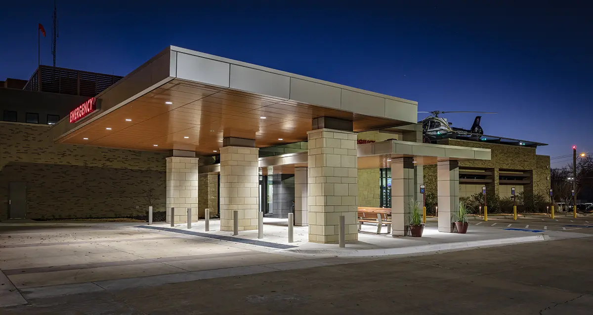 A well-lit hospital emergency entrance with stone pillars and a modern canopy at night. A helicopter is visible on the roof, adding urgency to the scene.