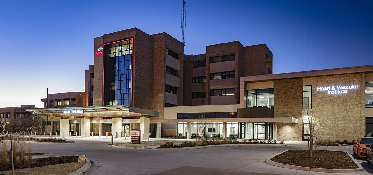 Modern hospital building illuminated at dusk, featuring prominent main entrance and "Heart & Vascular Institute" sign. A calm, welcoming atmosphere.