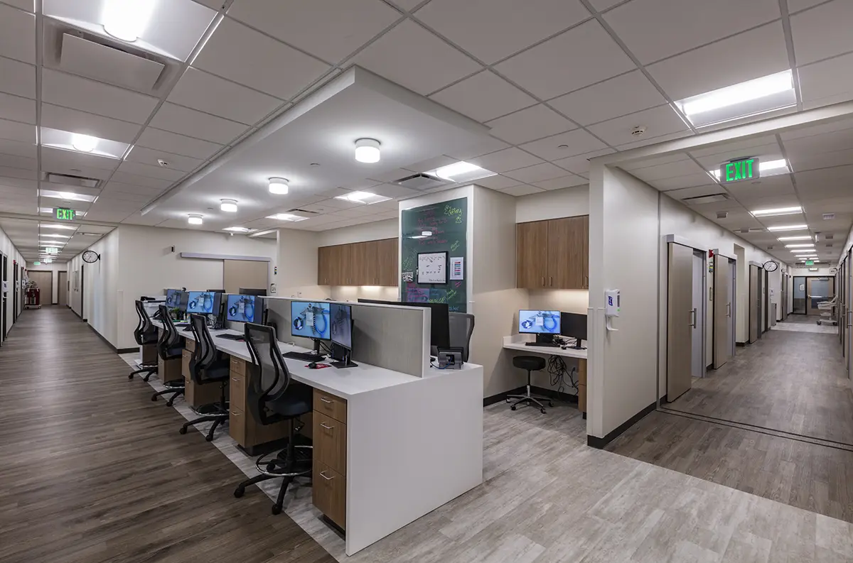 Modern medical office corridor with wood flooring, white walls, and ceiling lights. Desks with computers line the hallway, conveying a clean, professional atmosphere.