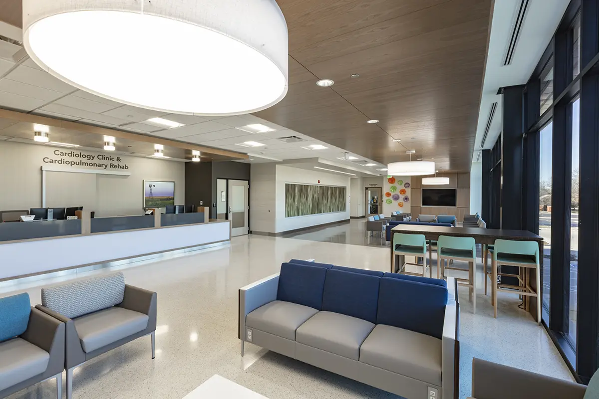 Modern clinic lobby with sleek design, featuring gray and blue couches, a high table with stools, large windows, and bright lighting. Calm and welcoming atmosphere.