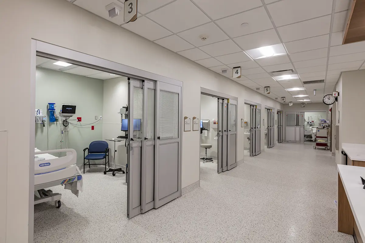 A hospital corridor with numbered patient rooms featuring sliding doors. Each room has medical equipment, a bed, and a chair. The atmosphere is sterile and organized.