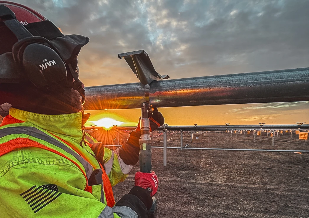 A construction worker in safety gear tightens a bolt on a steel beam at a solar farm during sunrise, conveying a sense of dedication and progress.
