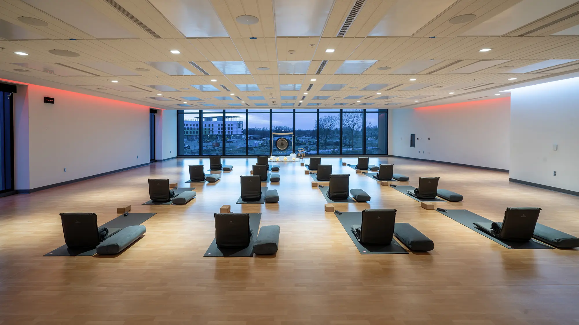 A spacious, modern yoga studio with large windows showing an urban view at dusk. Mats and cushions are neatly arranged, creating a calm, organized ambiance.