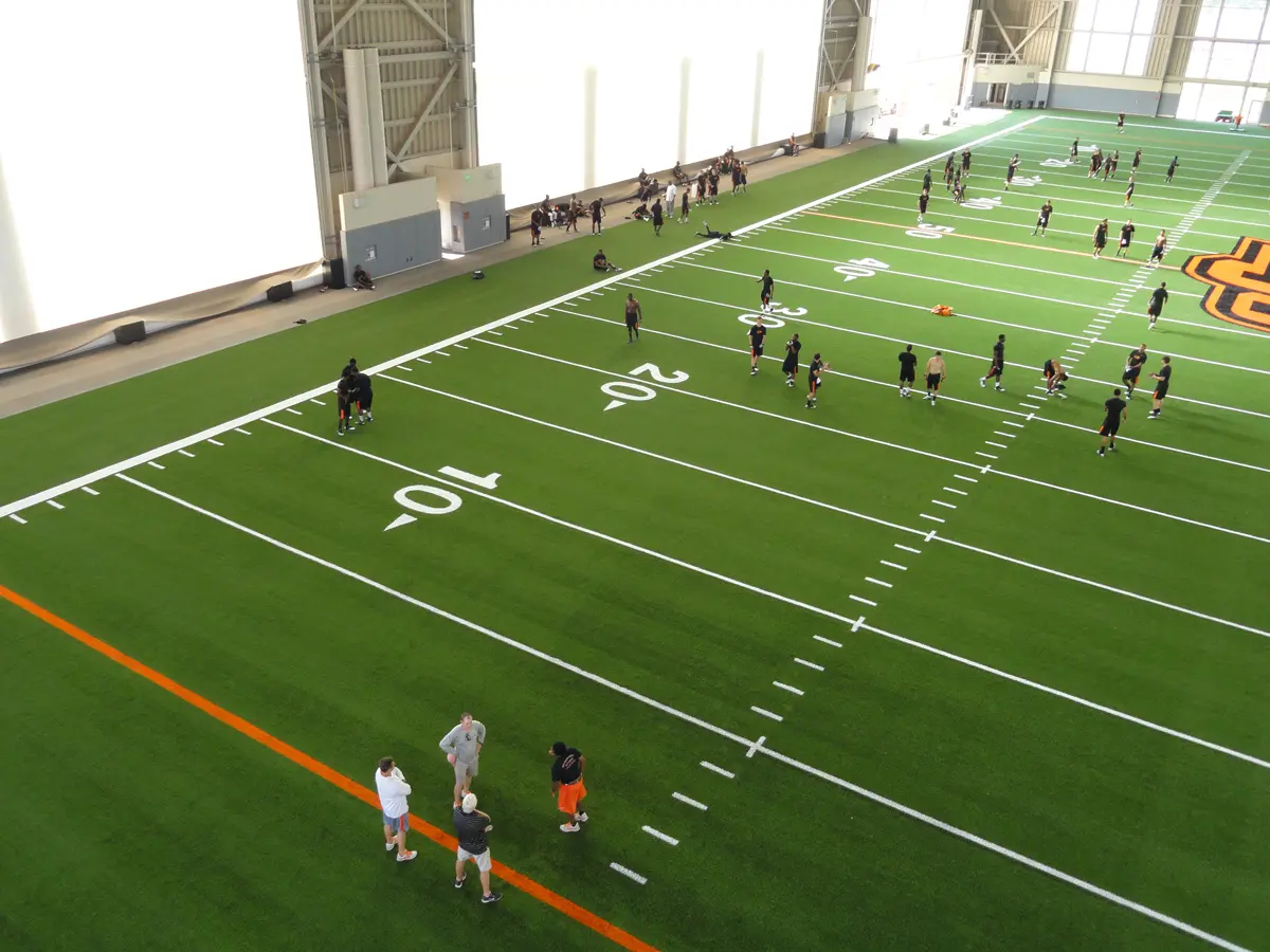 Indoor football practice field with players scattered. Coaches and athletes engage in training on green turf marked with yard lines. Bright, spacious ambience.