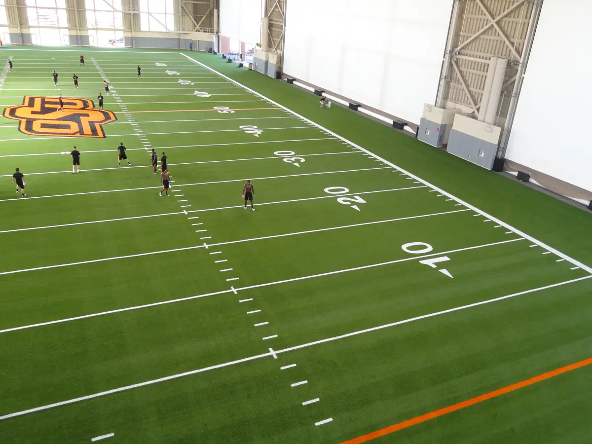 Indoor football field with green turf and white yard lines. A large orange and black logo is central. Several people are scattered, creating a casual, focused atmosphere.