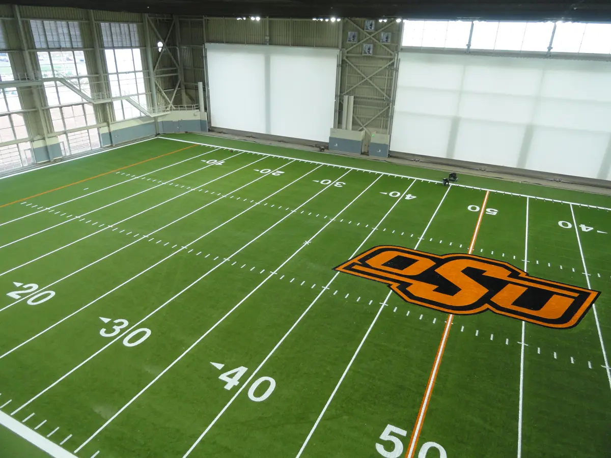 Indoor football field with green turf, marked with yard lines and an orange "OSU" logo at the 50-yard line. Bright, spacious facility.